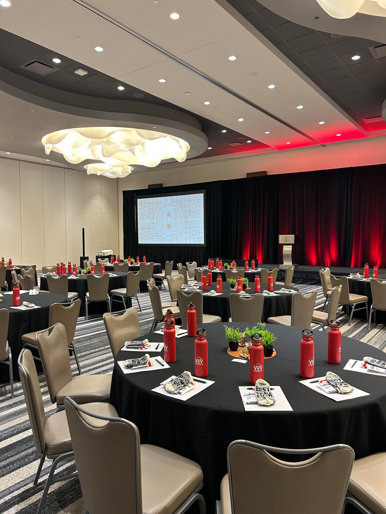 Ballroom set for a team meeting with black linens, branded red water bottles and a stage with red uplighting