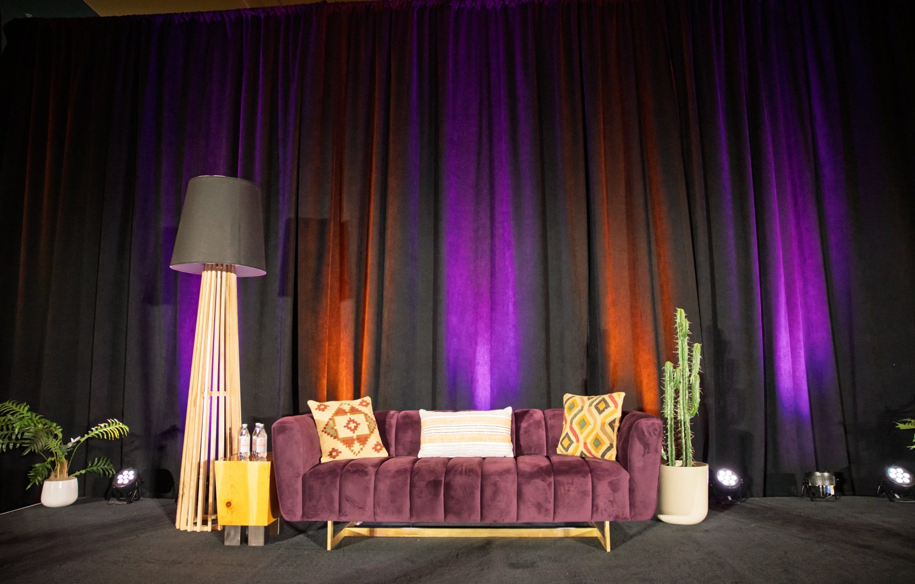 Stage set with a velvet sofa, wood floor lamp and cactus against lit drapes